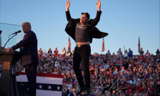 man wearing hat and black clothes jumps up as another man speaks at podium in front of crowd