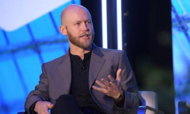 man with beard wearing grey suit and black shirt sits and gestures with hand